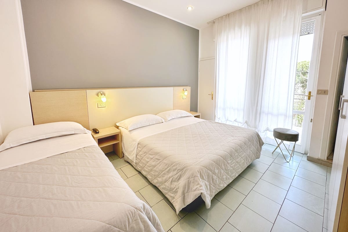 A hotel room with two beds, a nightstand with a lamp, a window with curtains, and a door leading to an outdoor area.