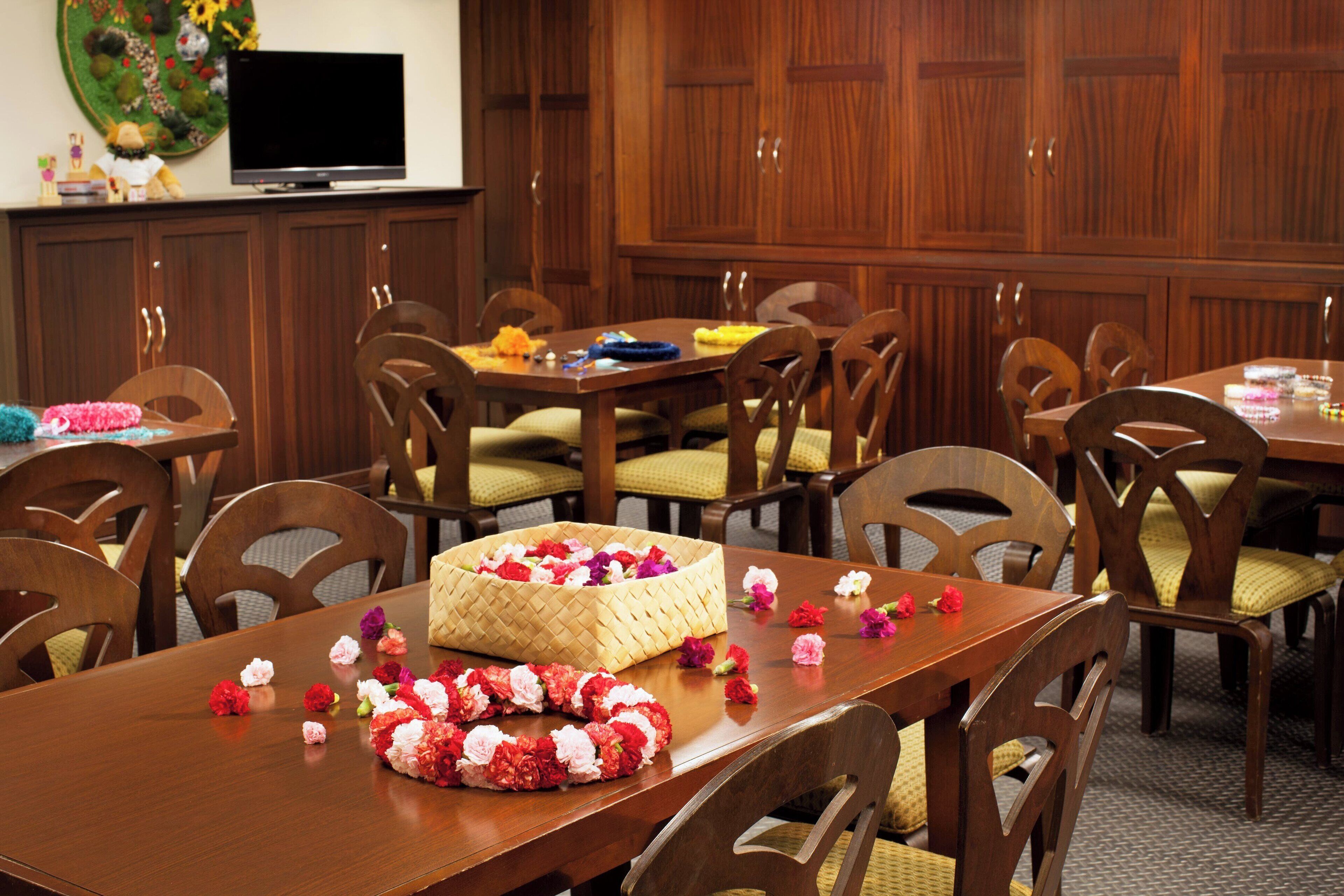 A dining area with wooden tables and chairs, a basket of flowers, and a television on the wall.