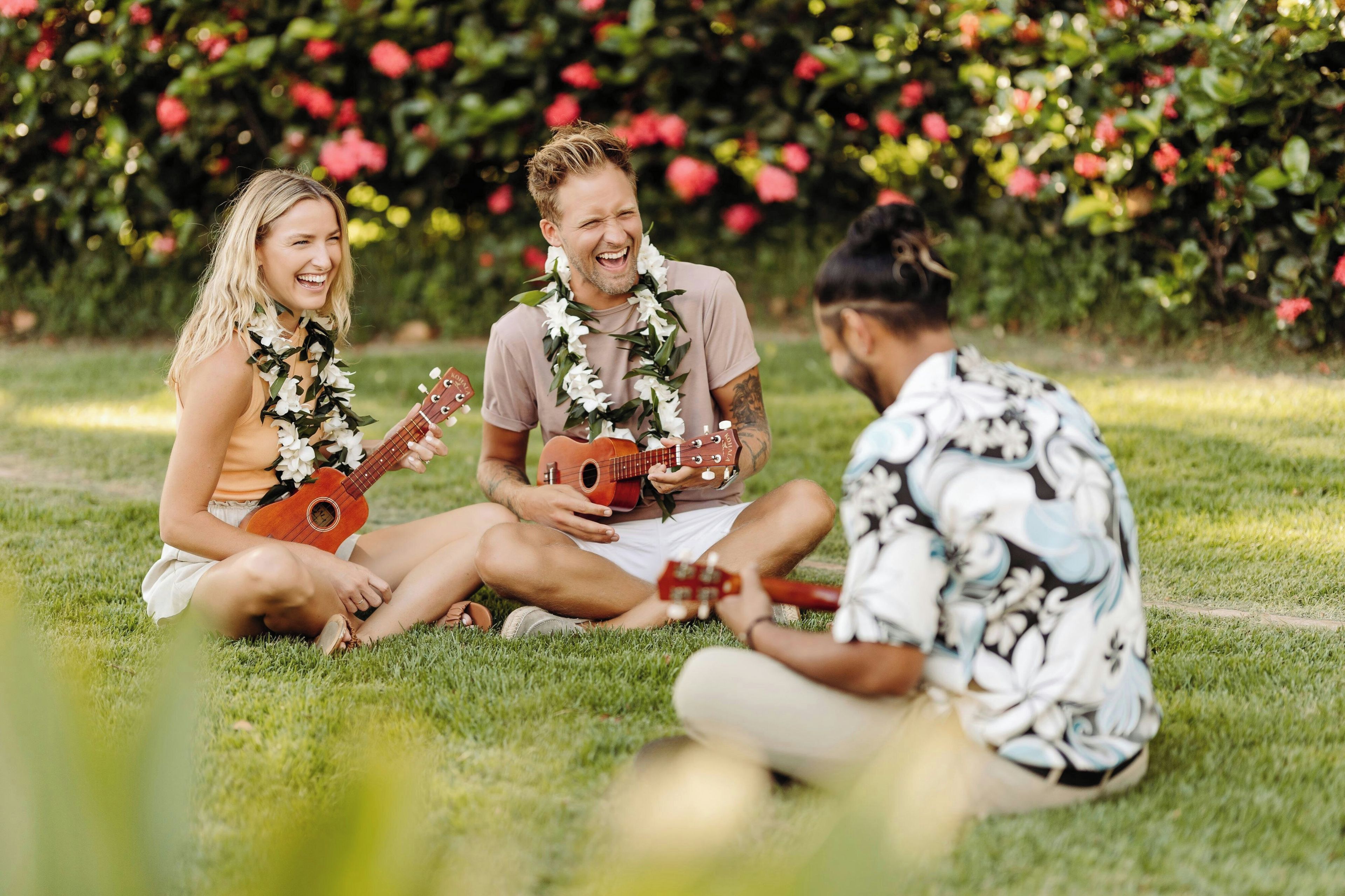 Three people sitting on grass with guitars, wearing leis, in a garden with red flowers.