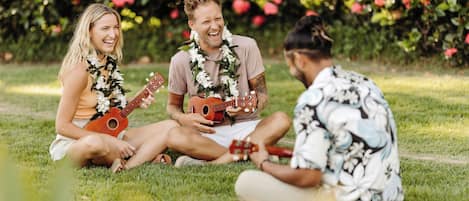 Three people sitting on grass with guitars, wearing leis, in a garden with red flowers.