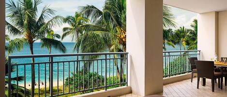A balcony with a view of the ocean and palm trees, a dining area with chairs, and a small table with a vase of flowers.
