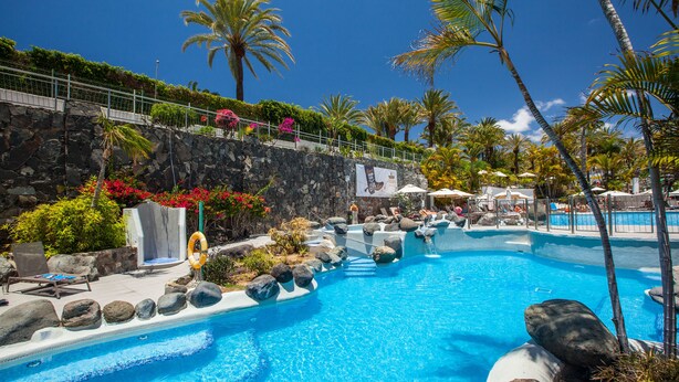 A swimming pool with a stone wall, palm trees, and lounge chairs. - Abora Catarina by Lopesan Hotels - All Inclusive (San Bartolomé de Tirajana)