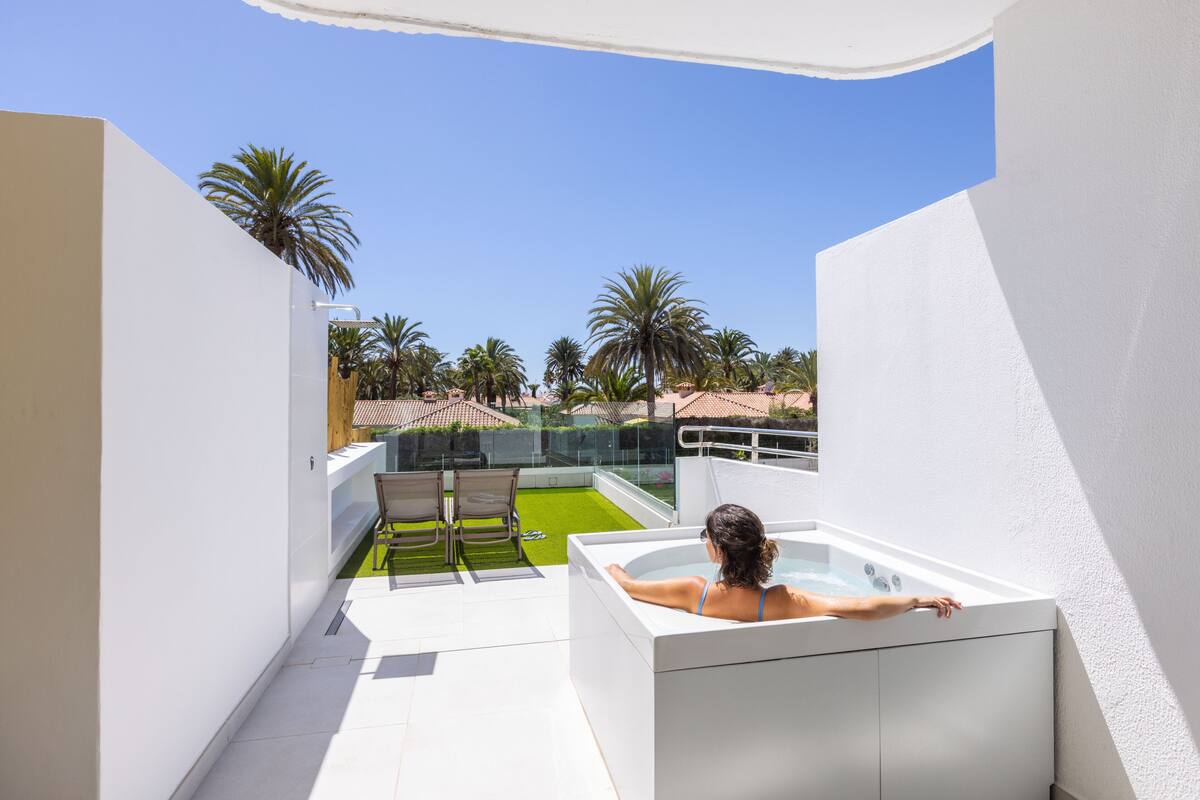 A woman relaxing in a white bathtub on a balcony overlooking a pool and palm trees.