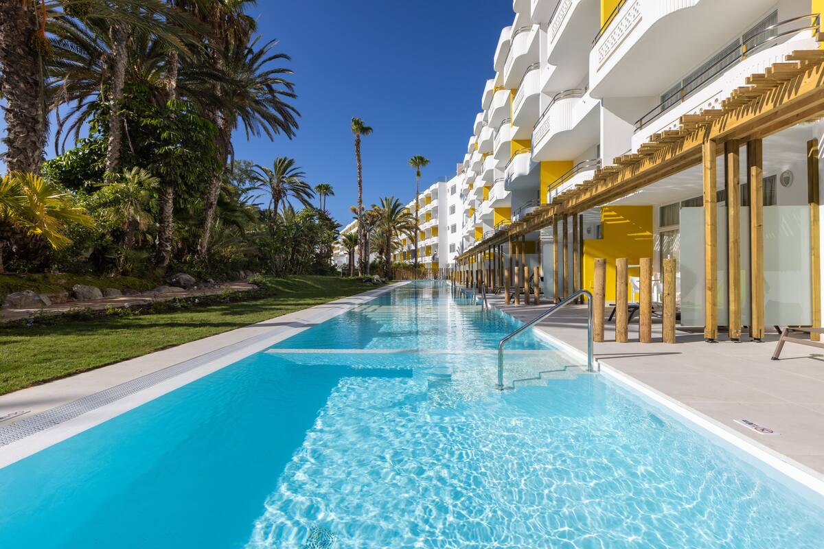 A long swimming pool with clear blue water, surrounded by palm trees and a modern building with balconies.