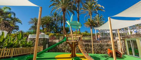 Un parque infantil con toboganes y estructuras para escalar, rodeado de palmeras y un cielo azul despejado.
