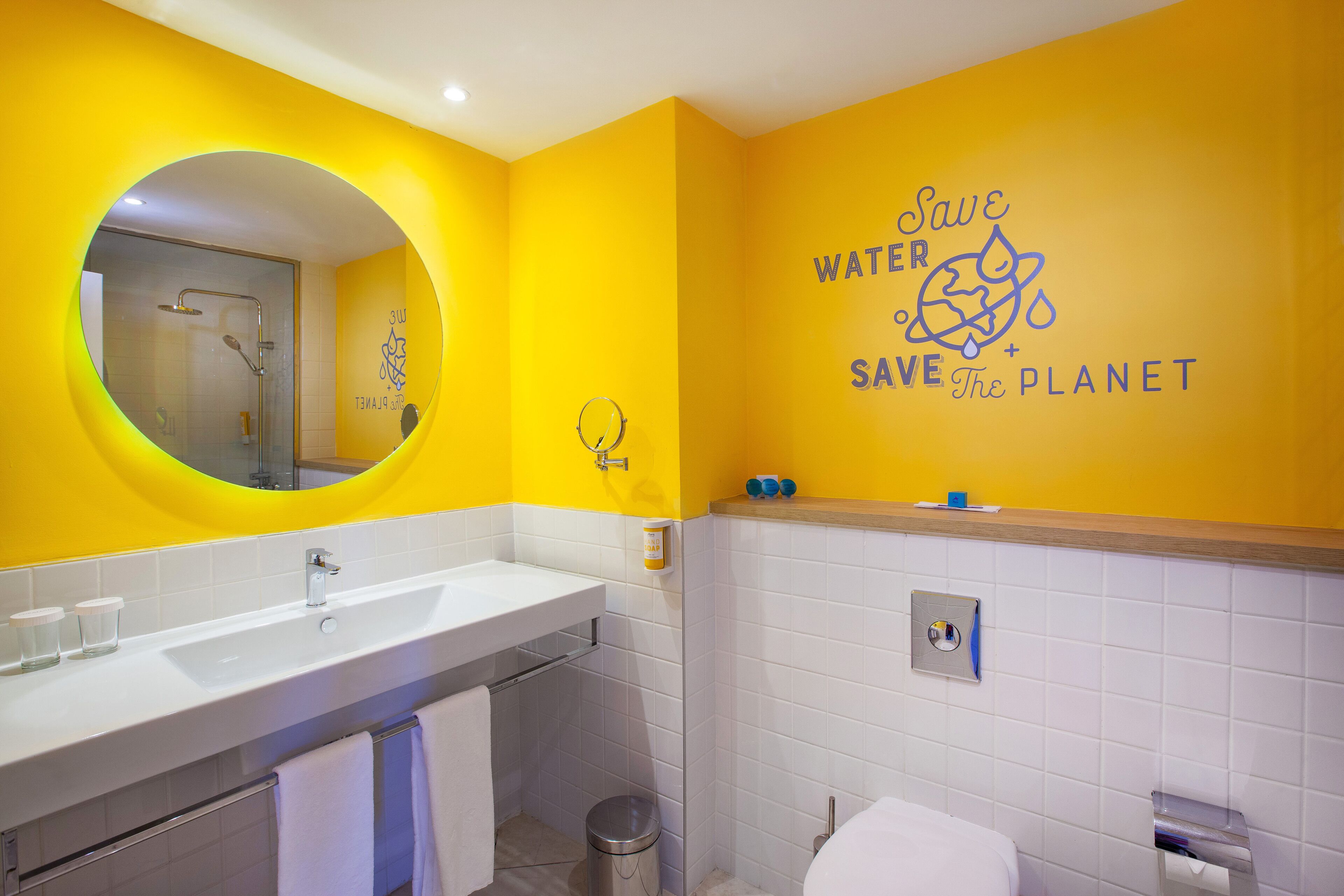 A bathroom with a yellow wall, a white sink, a mirror, and a toilet. There is a sign on the wall that says 'Save Water Save the Planet'.