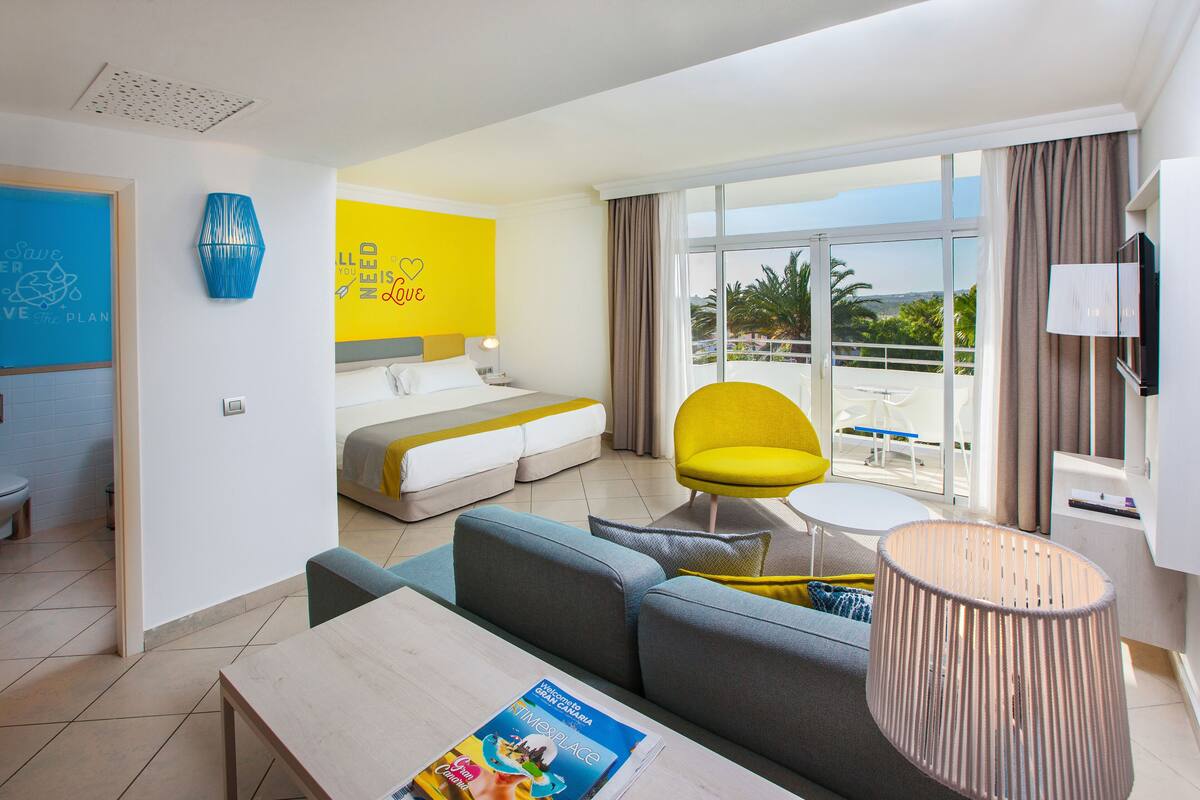 A hotel room with a bed, a sofa, a dining table, and a balcony with a view.