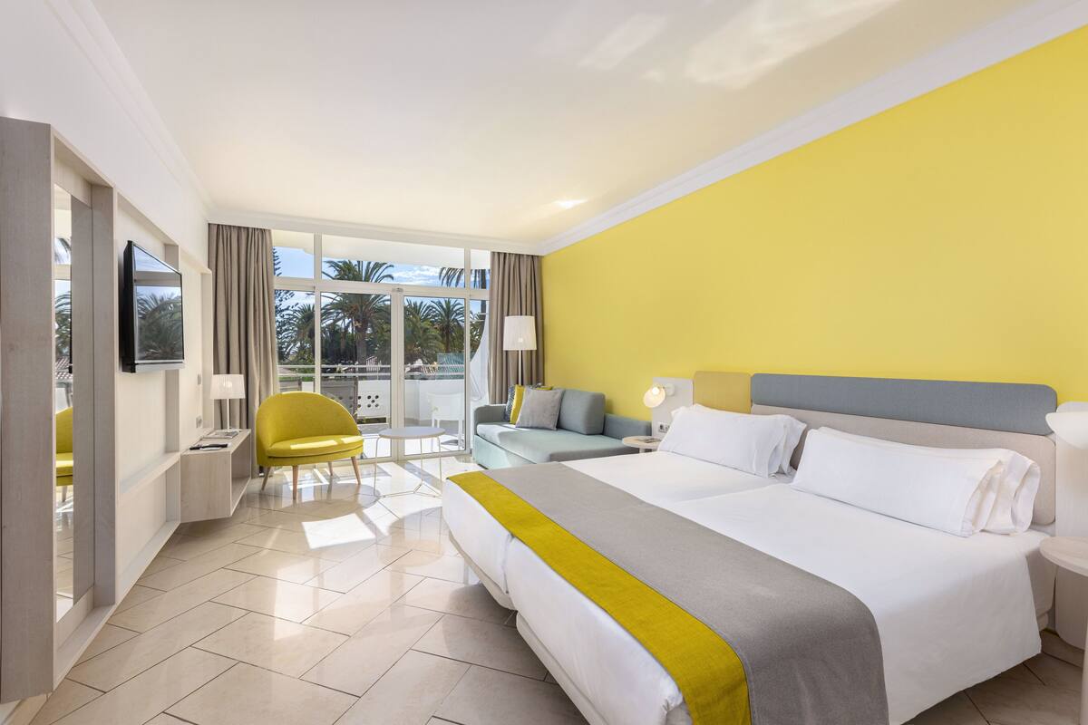 A hotel room with a large bed, a sofa, a chair, a TV, and a balcony with a view of palm trees.