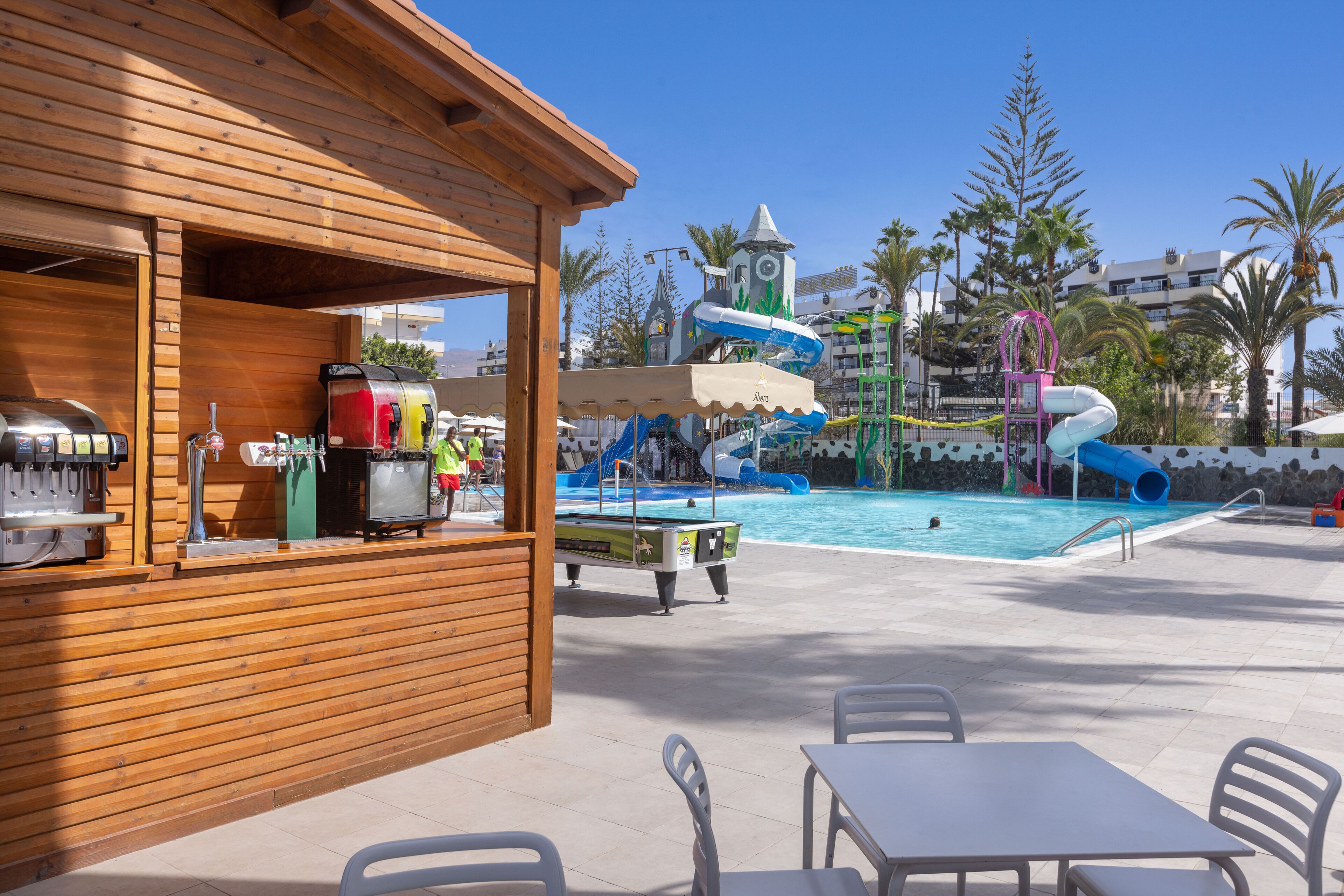 A pool area with a wooden bar, tables, and chairs, and a water slide in the background.