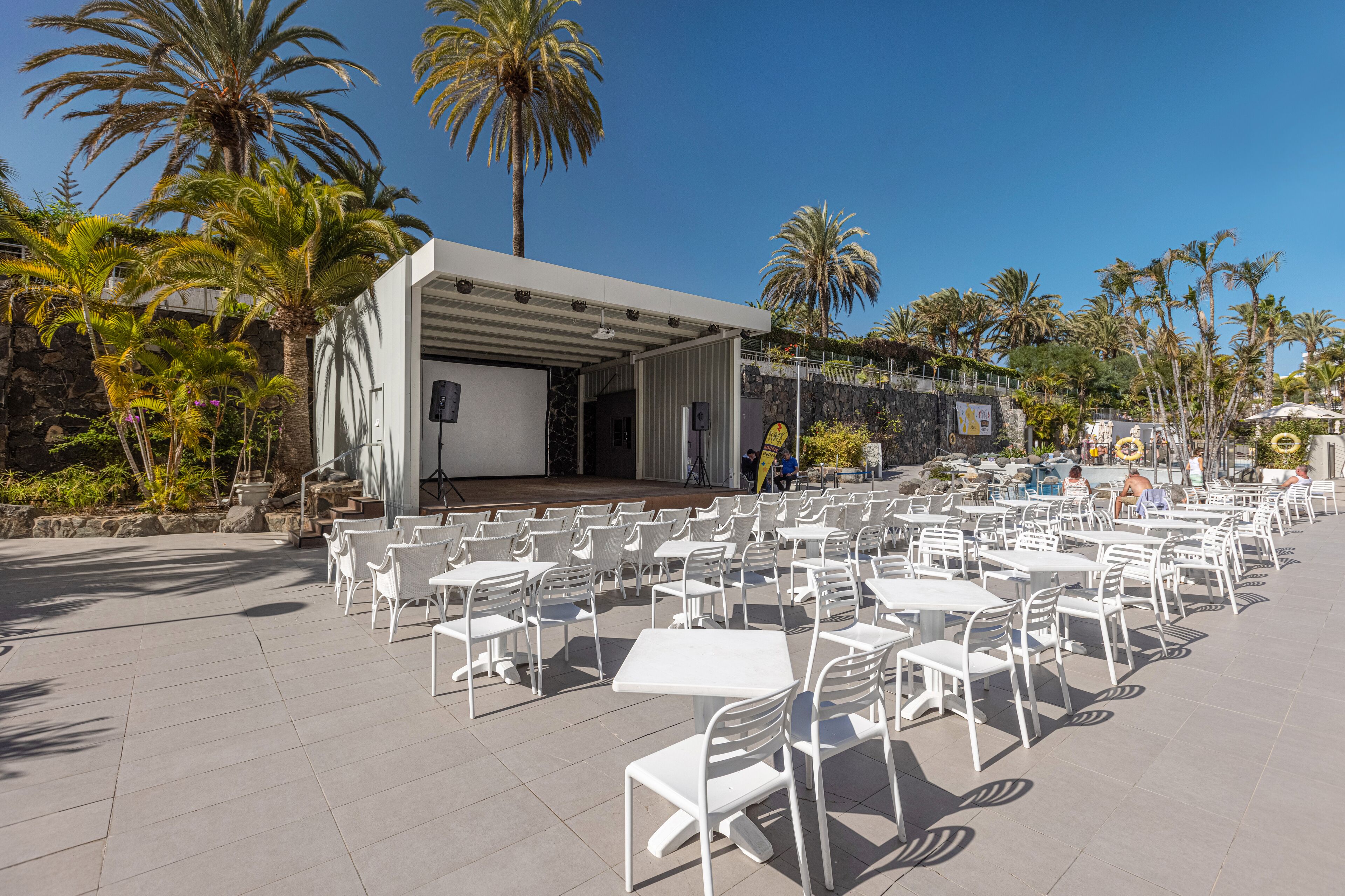 An outdoor seating area with white tables and chairs, a stage, and palm trees.