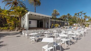 An outdoor seating area with white tables and chairs, a stage, and palm trees.