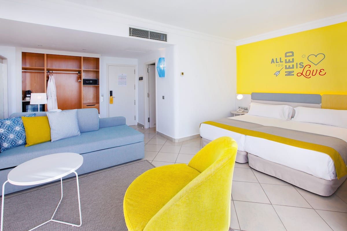 A modern hotel room with a bed, a sofa, a yellow armchair, and a small white table.