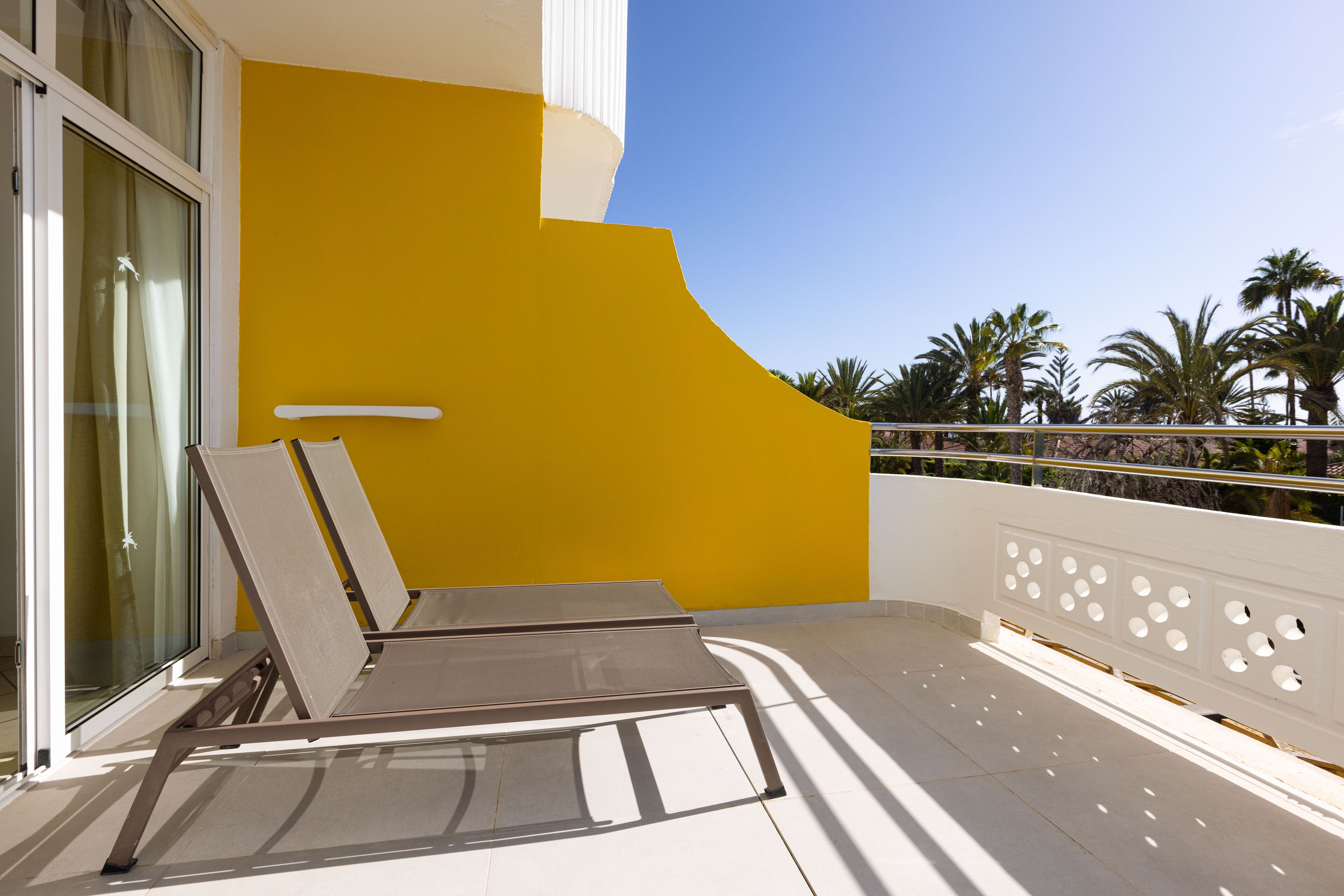 Ein Balkon mit gelber Wand, weißer Brüstung und Blick auf Palmen.