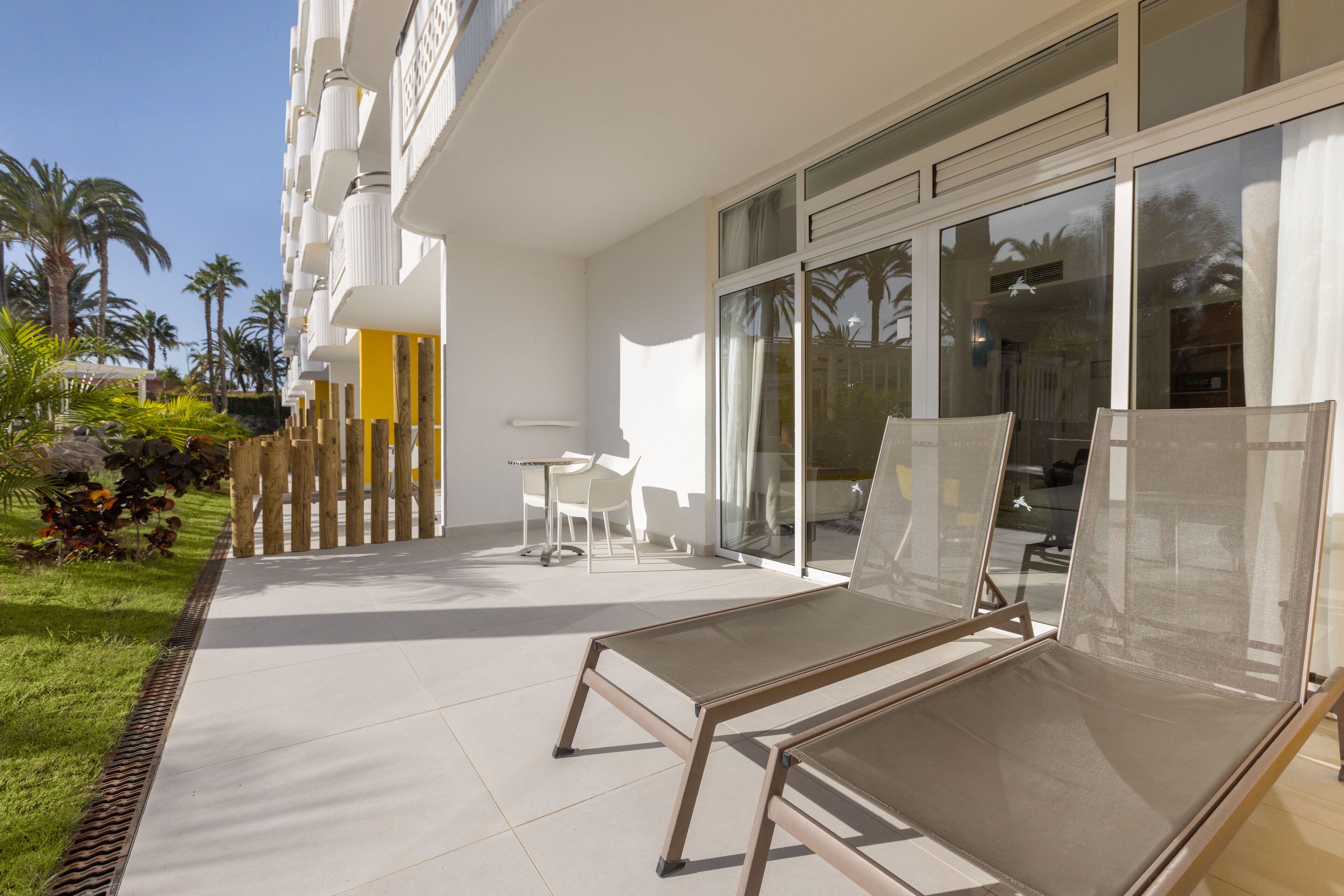 A modern hotel exterior with a covered outdoor seating area, a pool, and a view of palm trees.