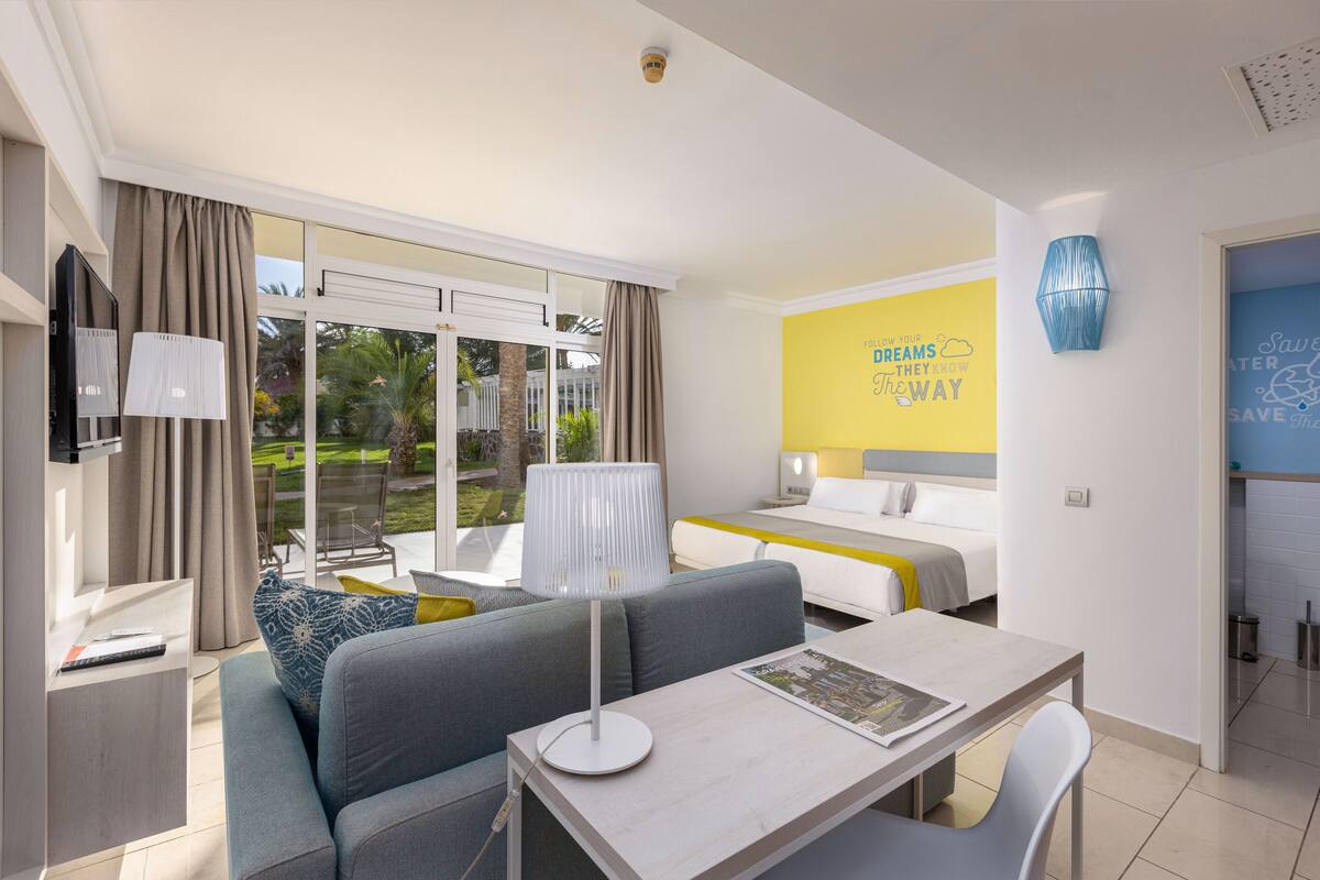 A modern hotel room with a sofa, a dining table, a bed, and a large window offering a view of the outdoors.