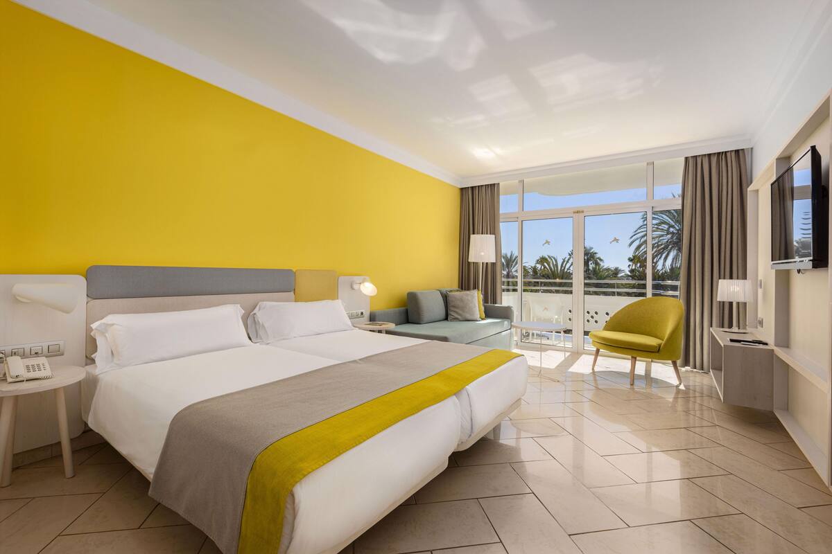 A hotel room with a large bed, a sofa, a chair, a TV, and a balcony with a view of palm trees.