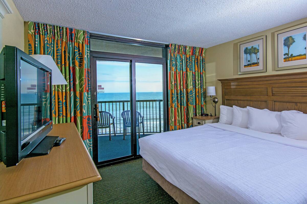 A hotel room with a bed, a desk with a television, a view of the ocean through a sliding door, and two framed pictures on the wall.
