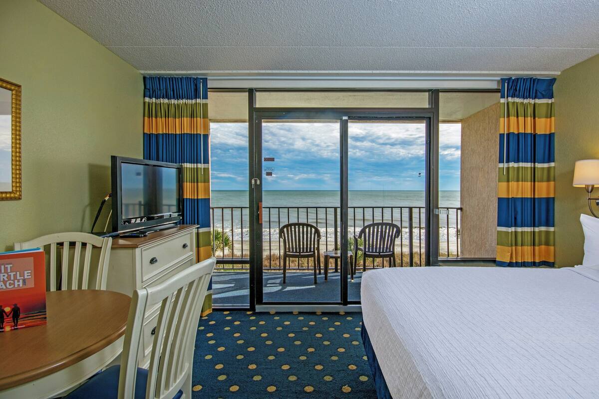 A hotel room with a bed, a TV, a desk, and a chair. There is a balcony with a view of the ocean.