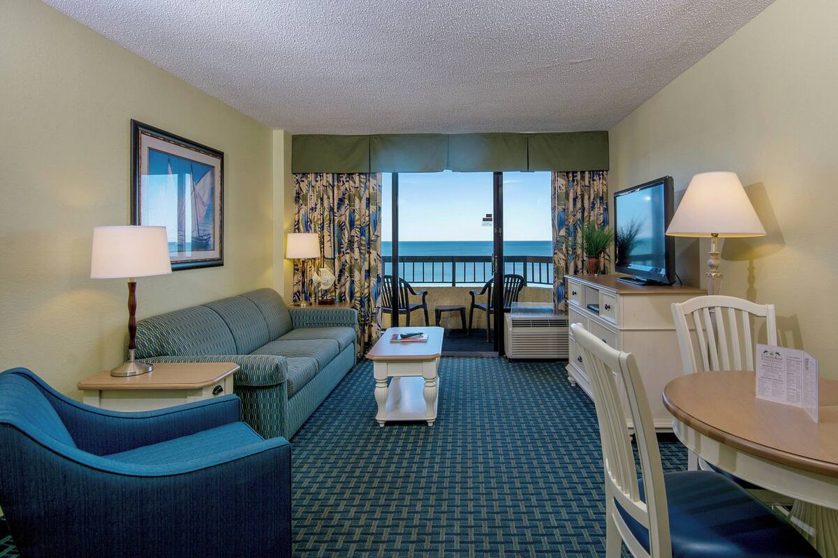 A hotel room with a balcony, a sofa, a dining table, and a TV.