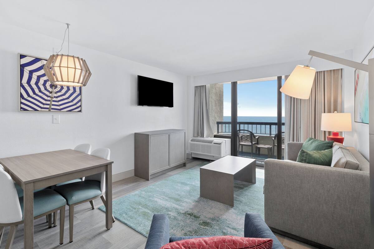 A modern hotel room with a dining area, a sofa, a TV, and a view of the sea.