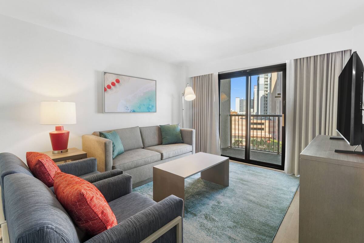 A modern hotel room with a grey sofa, a red lamp, a flat-screen TV, and a large window with a city view.