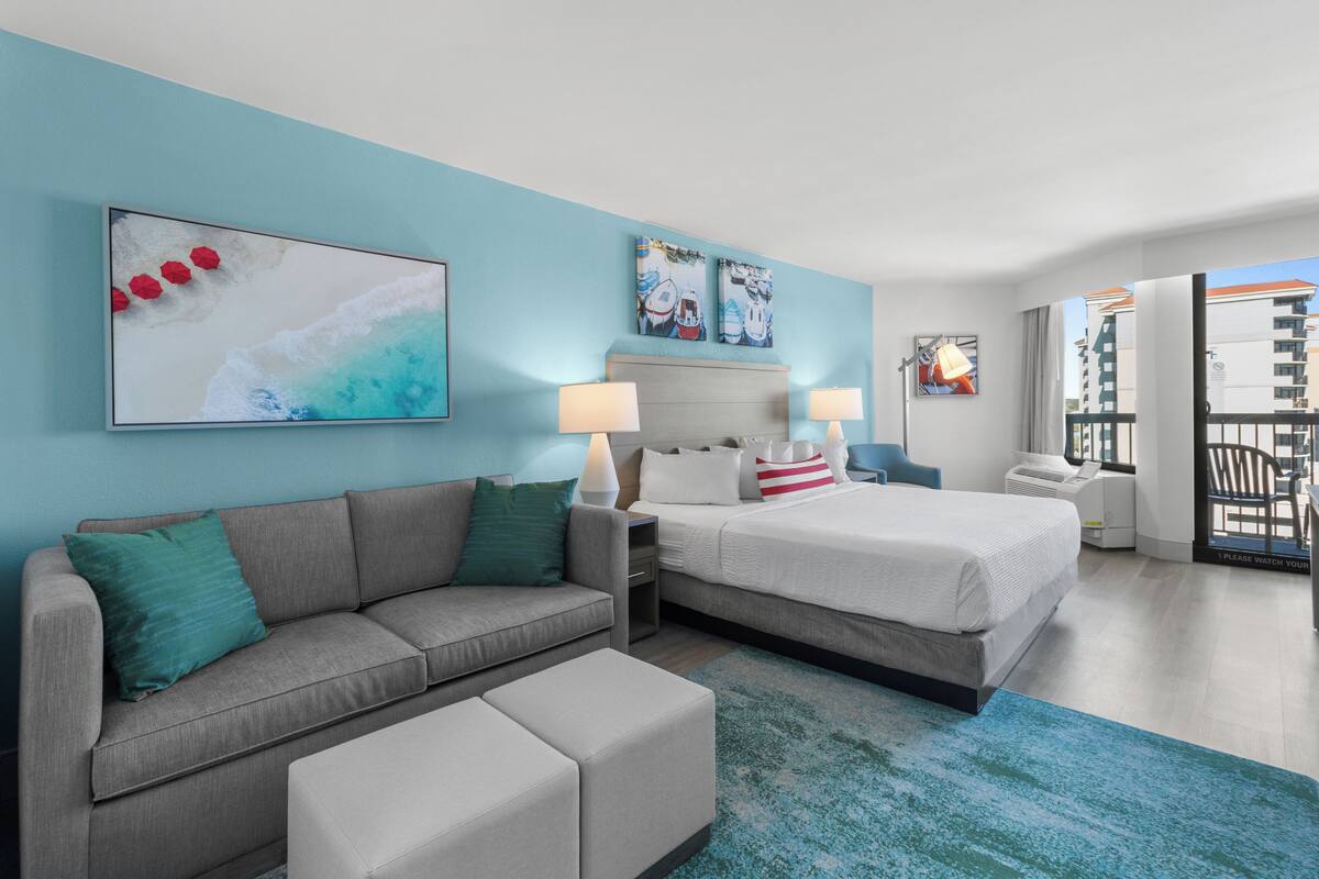 A modern hotel room with a grey sofa, a bed with white bedding, a blue accent wall, and a large window offering a view of the city.