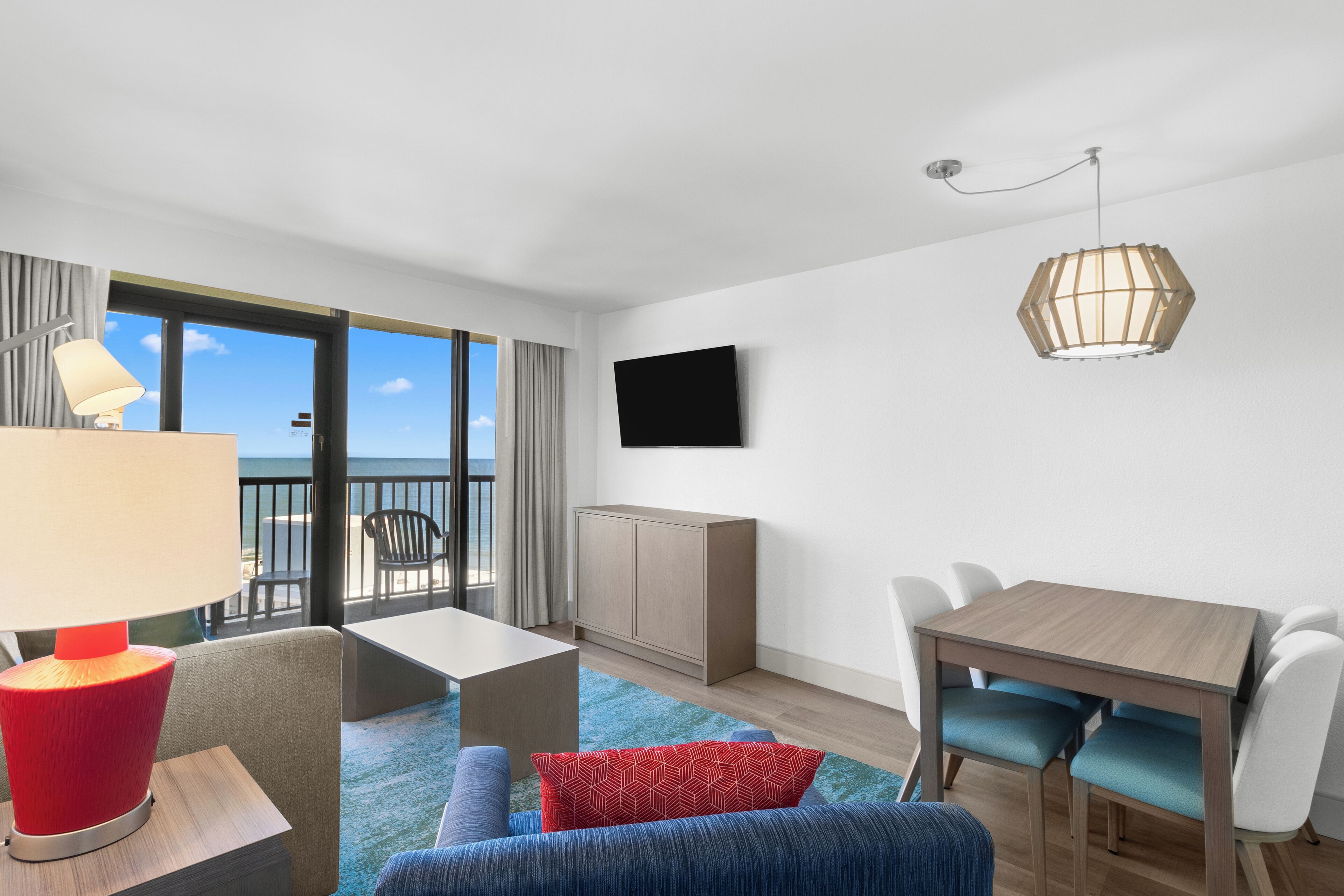 A modern hotel room with a balcony, a flat-screen TV, a wooden dining table with chairs, a red lamp, and a blue sofa.