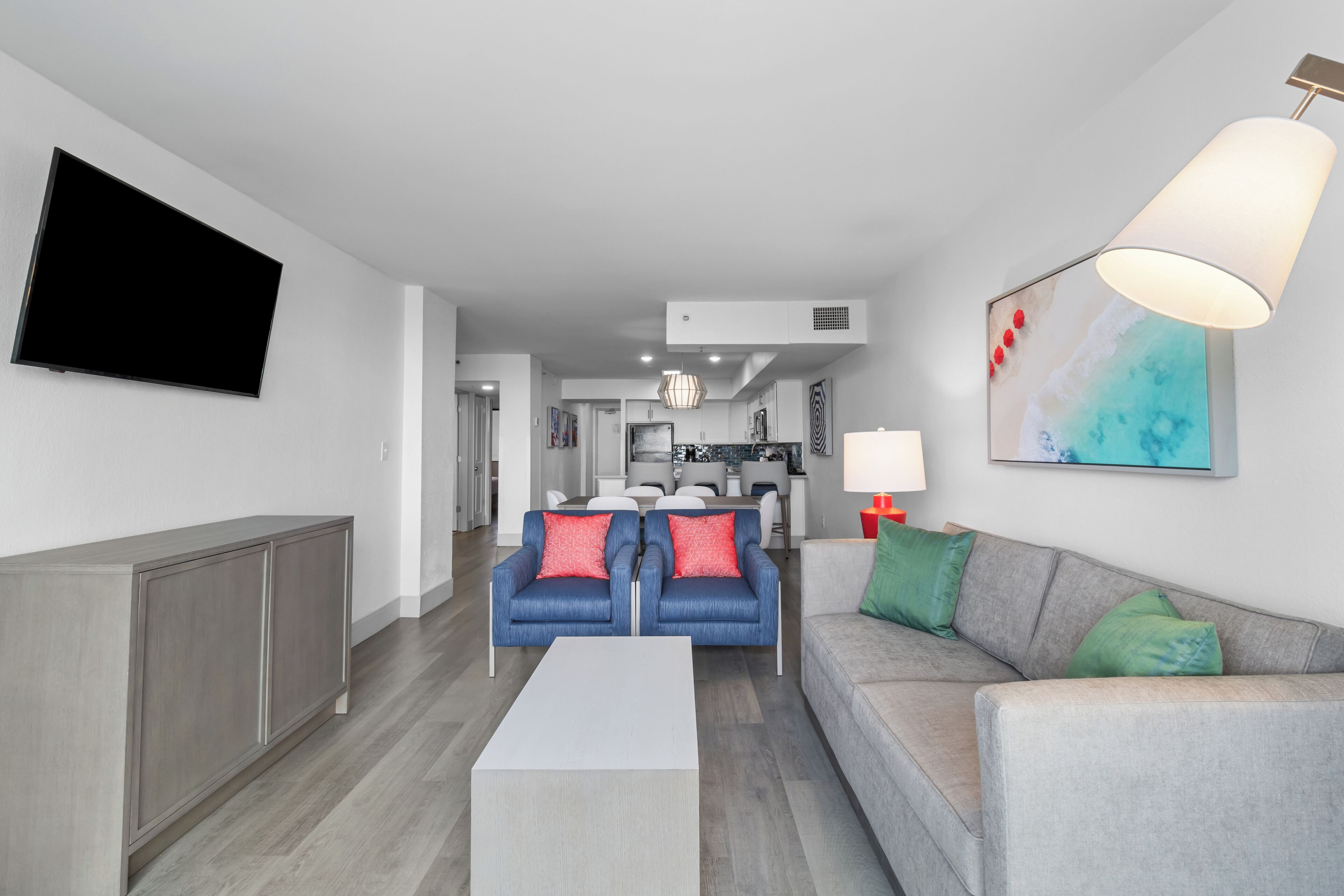 A modern living room with a grey sofa, a blue armchair, and a white coffee table. There is a flat-screen TV mounted on the wall and a lamp on the right side.