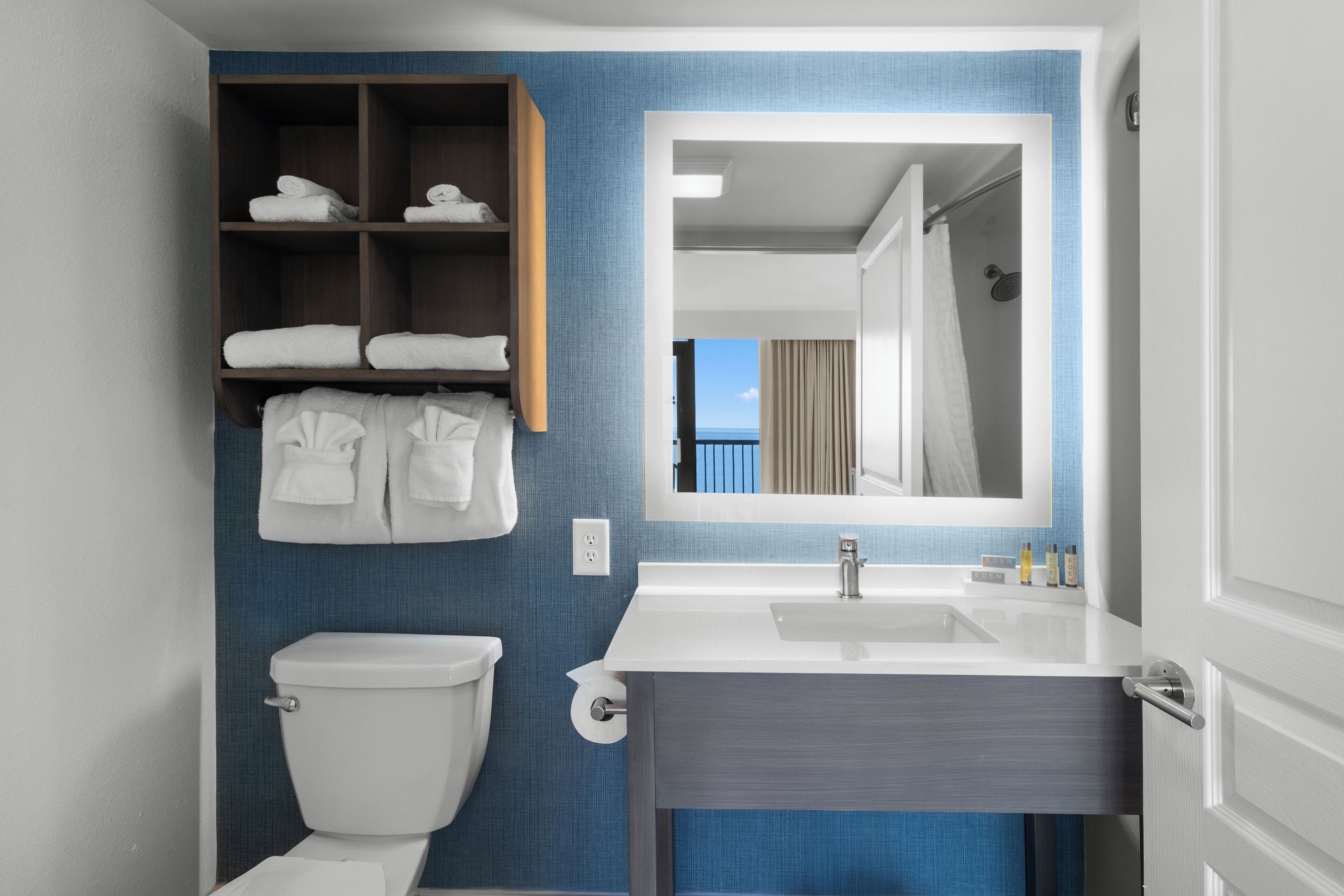 A modern bathroom with a white toilet, a built-in shelf with towels, a large mirror, and a sink with a vanity.