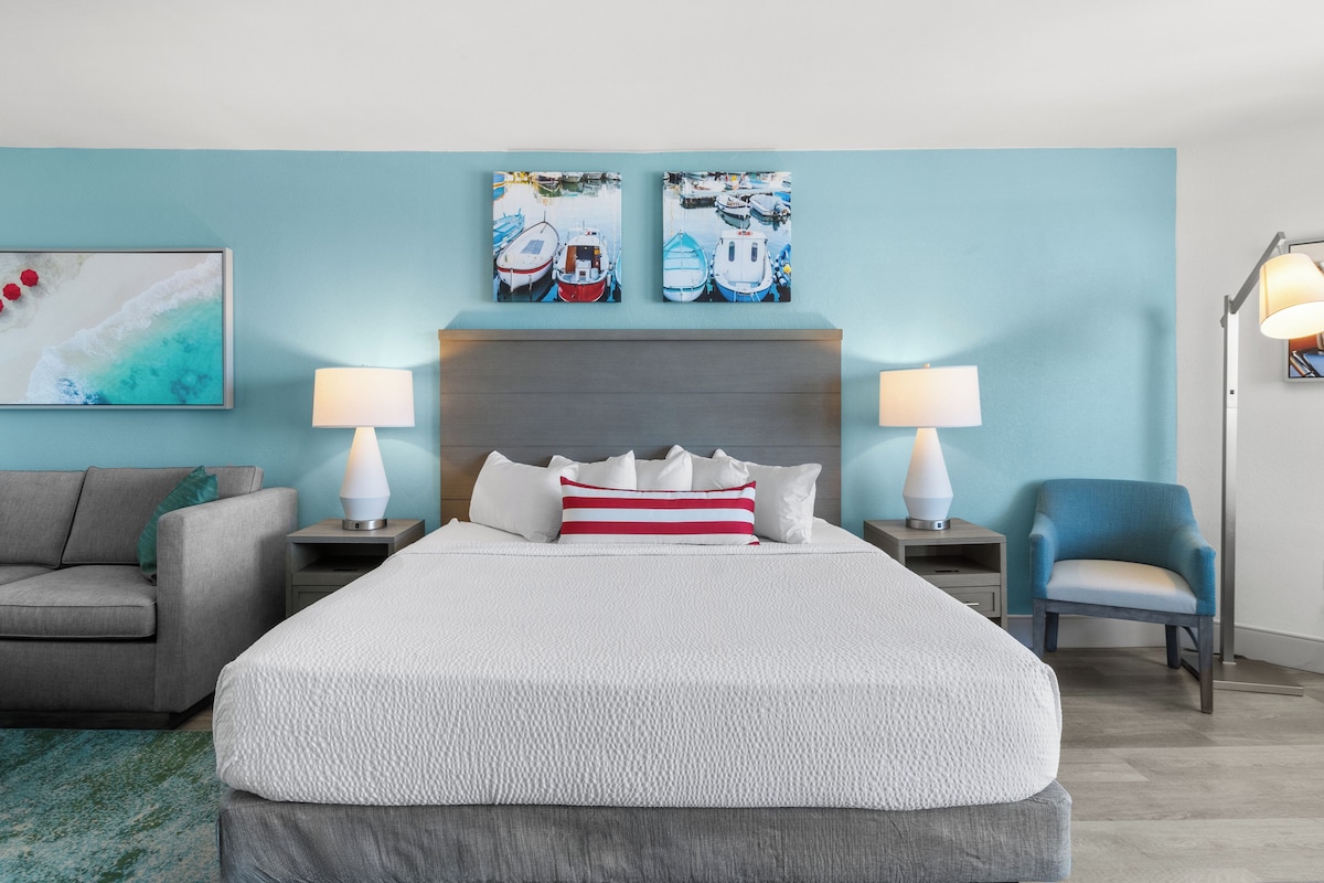 A hotel room with a large bed, two bedside tables with lamps, a grey sofa, a blue armchair, and artwork on the walls.