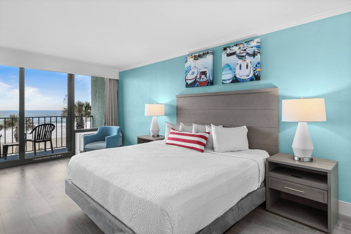 A modern hotel room with a large bed, two bedside tables with lamps, a view of the beach, and two wall-mounted artworks.