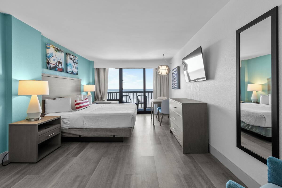 A modern hotel room with a large bed, a desk, a chair, a dresser, and a large mirror.