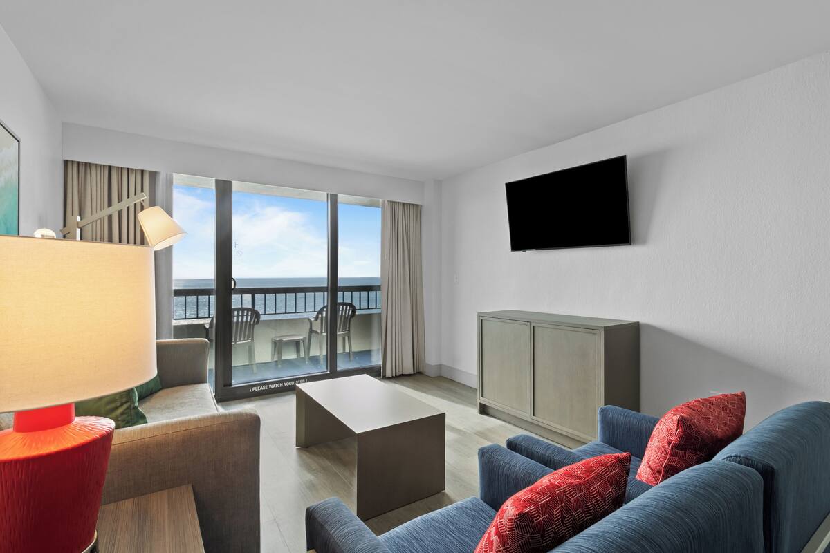 A modern hotel room with a sofa, a small table, a TV mounted on the wall, and a balcony with a view.