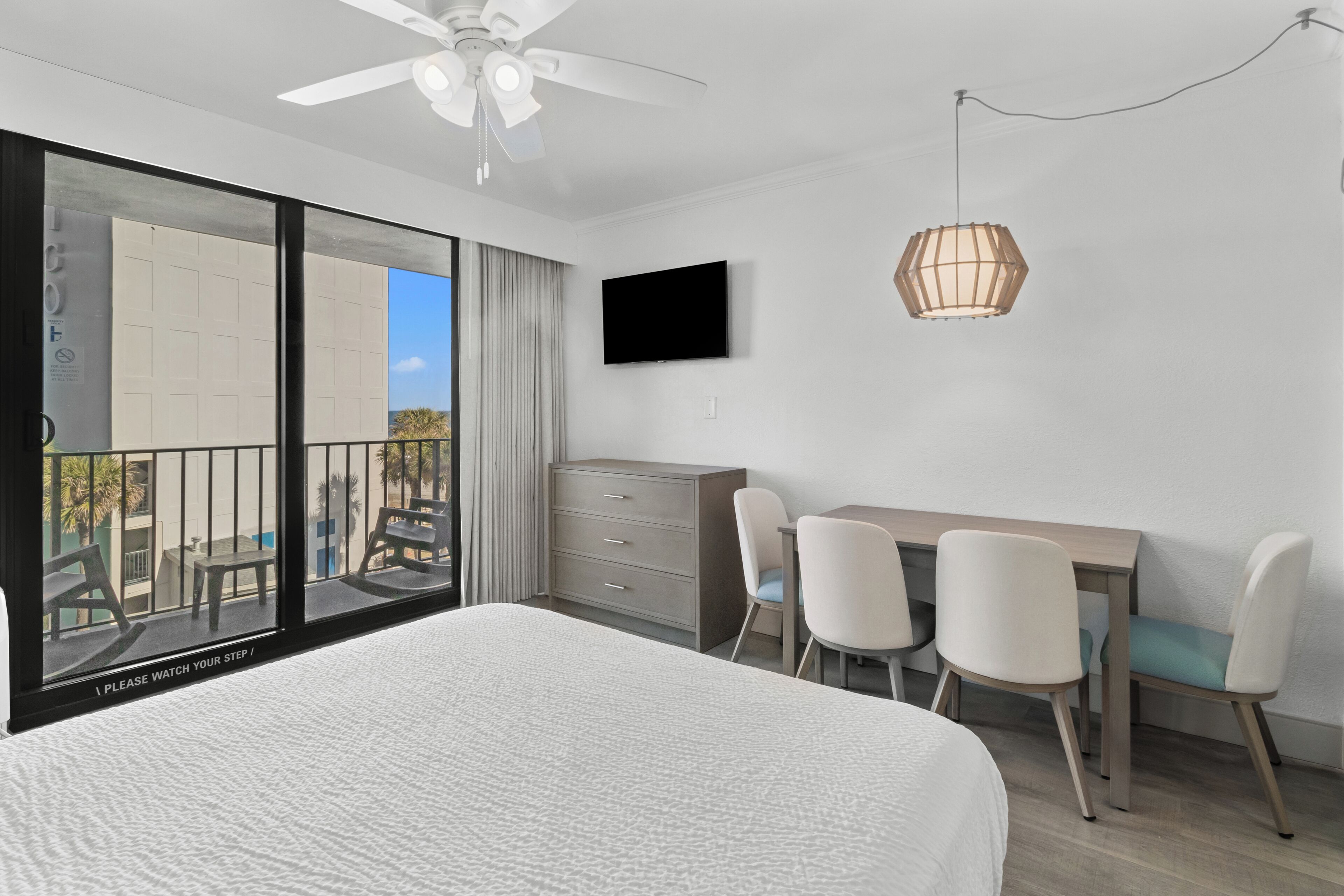 A modern hotel room with a large bed, a desk with chairs, a television, and a balcony with a view.