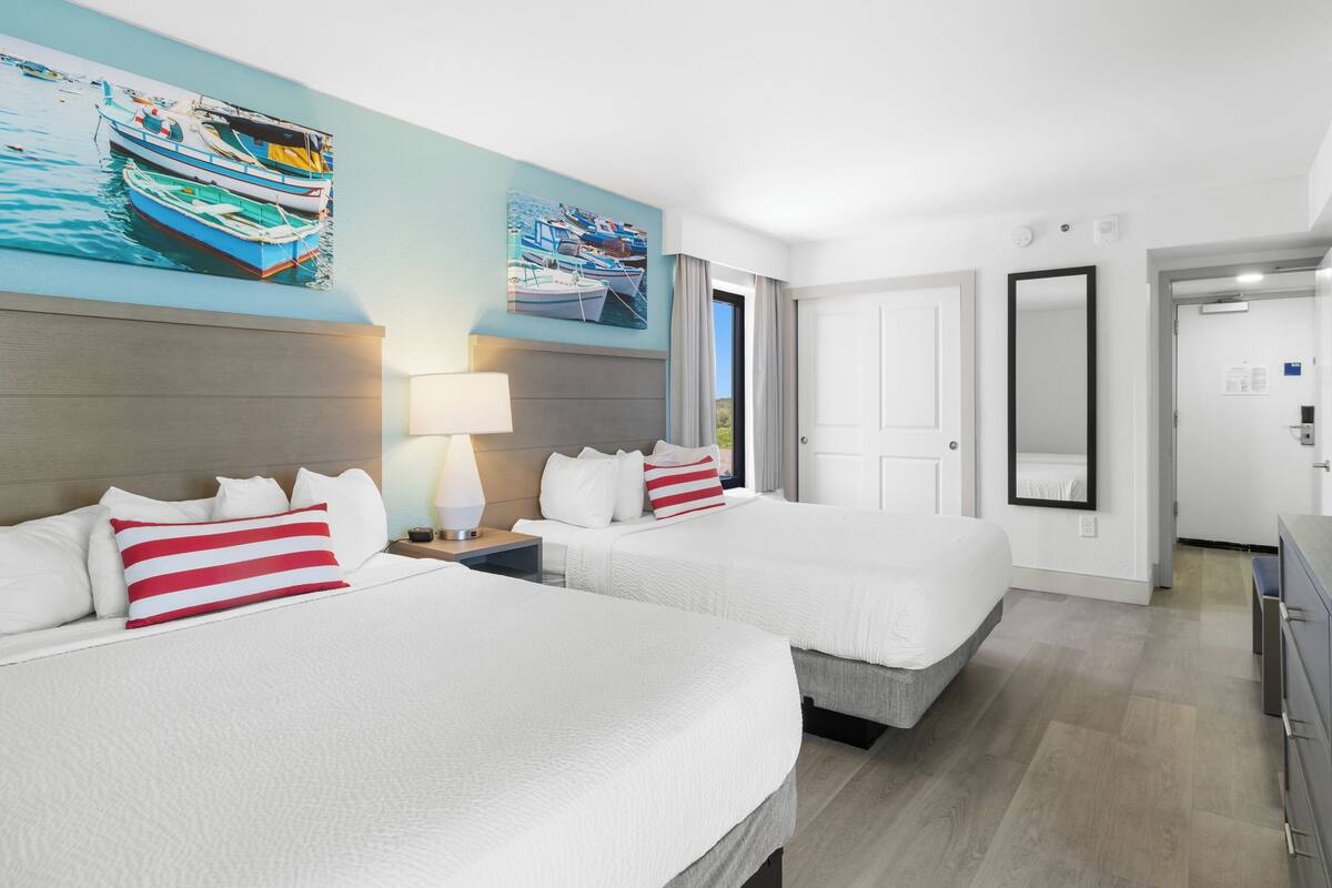 A hotel room with two beds, a large painting of boats on the wall, a door, and a mirror.