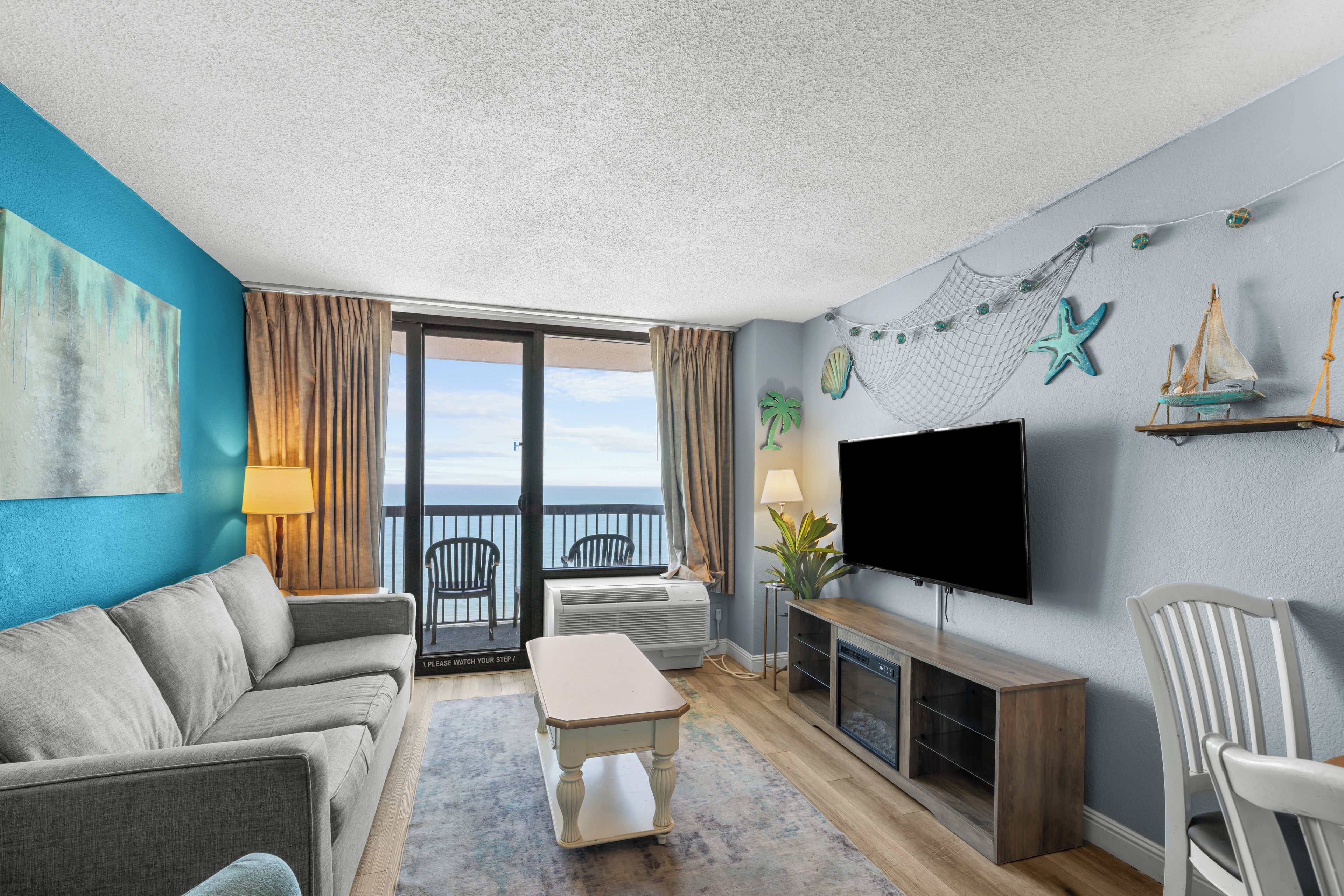 A living room with a sofa, a coffee table, a TV, and a balcony with a view of the sea.