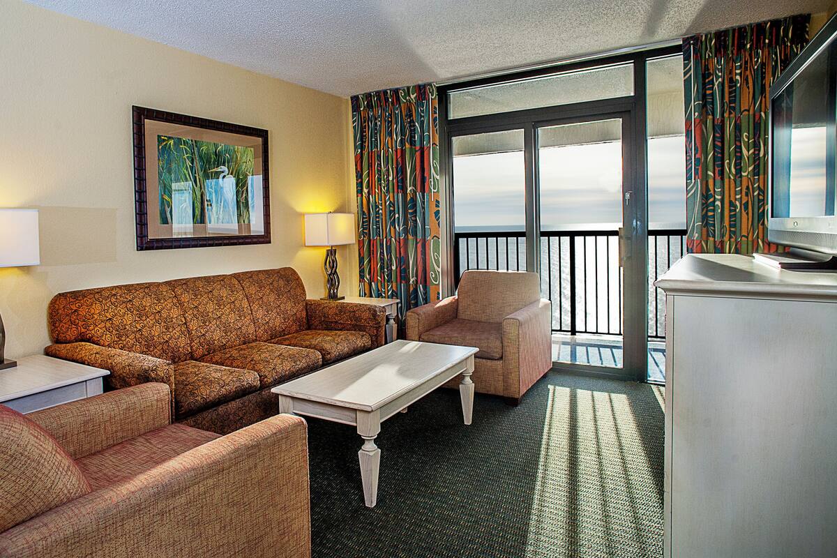 A hotel room with a sofa, armchair, and a small table. There is a painting on the wall and a television on a stand.