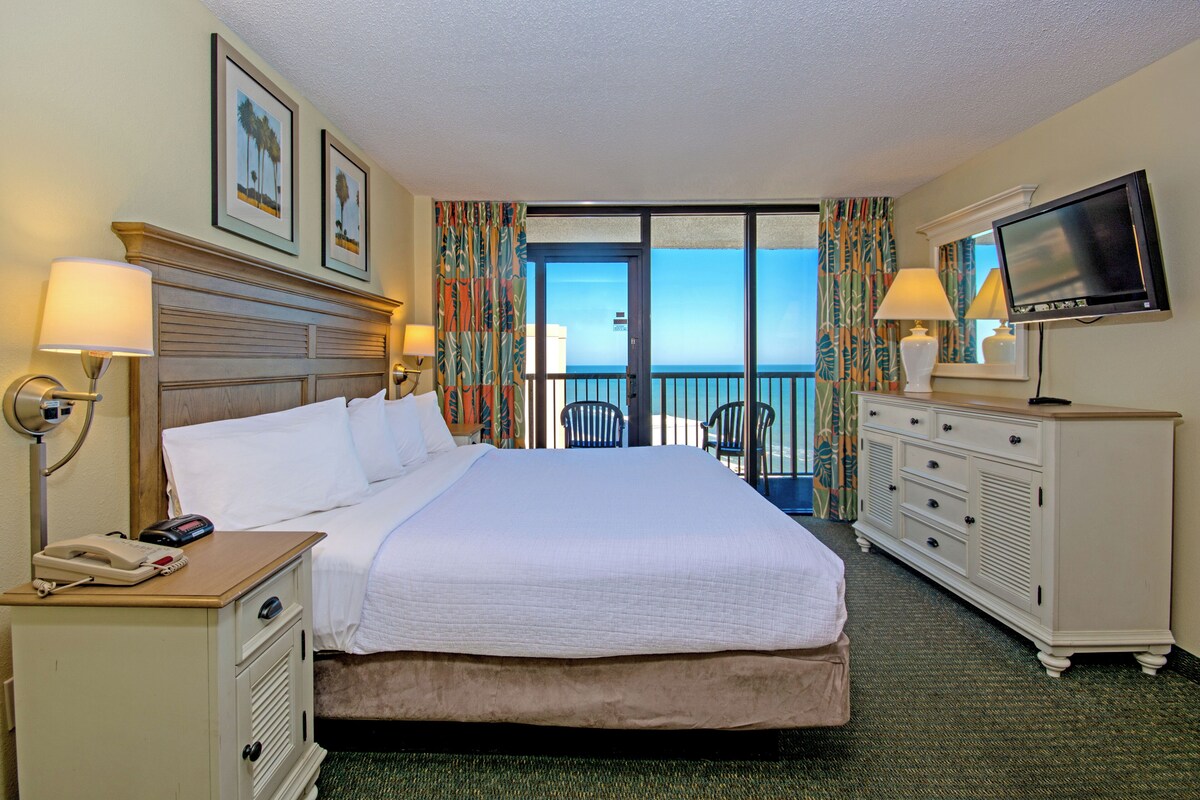 Una habitación de hotel con cama, lámparas de noche, televisor, cómoda y vista al océano.