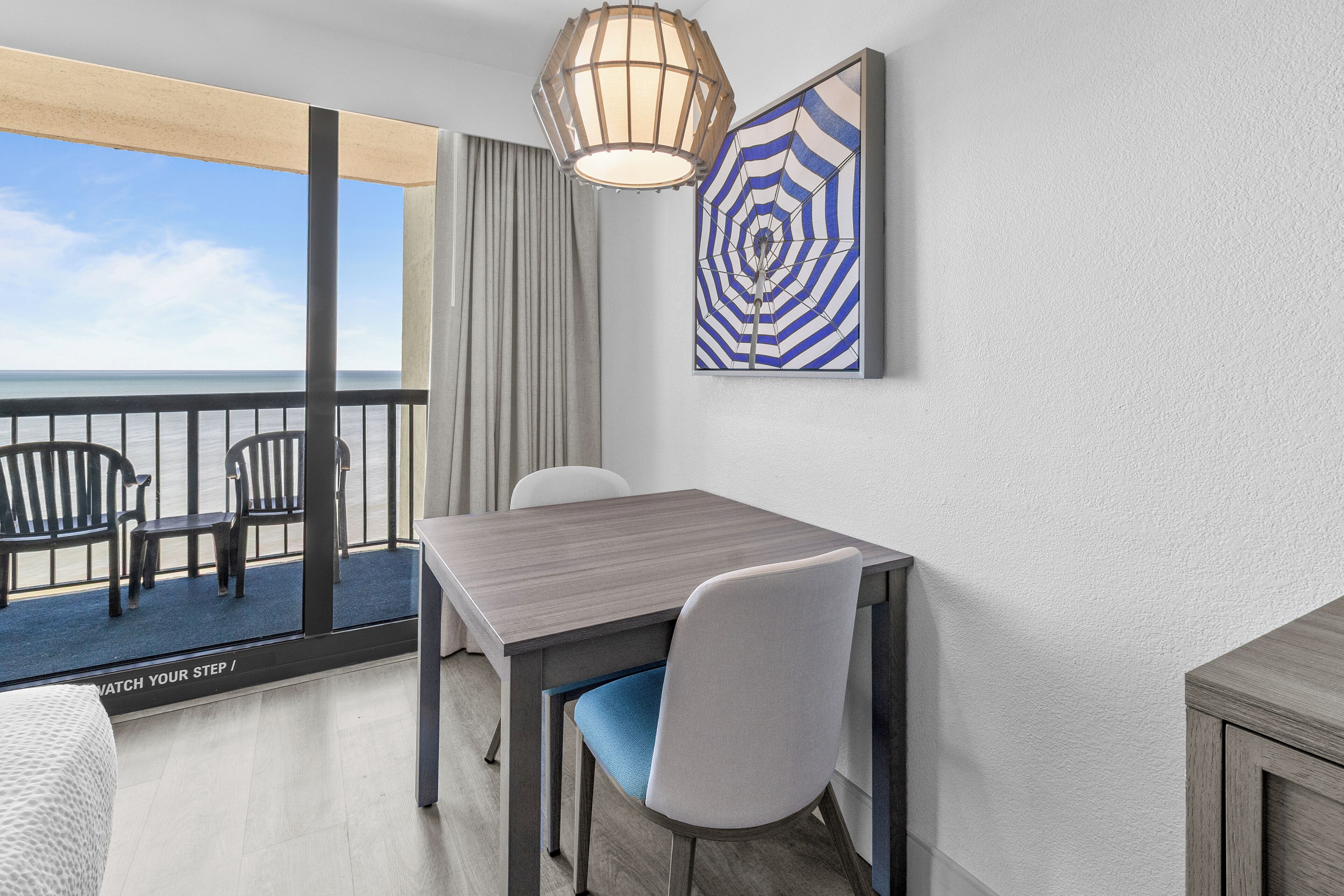 A balcony with a view of the ocean, a table with chairs, and a piece of abstract art on the wall.