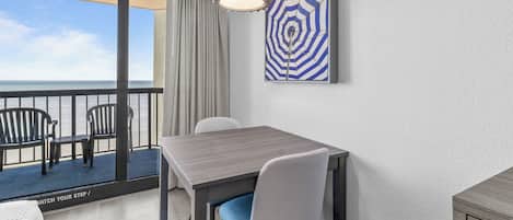 A balcony with a view of the ocean, a table with chairs, and a piece of abstract art on the wall.