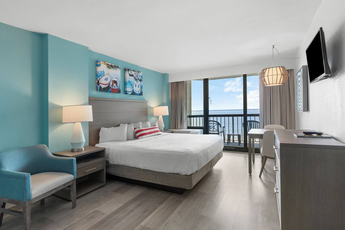 A modern hotel room with a large bed, two bedside tables, a desk, and a chair. There are two paintings on the wall, a TV mounted on the wall, and a balcony with a view of the ocean.