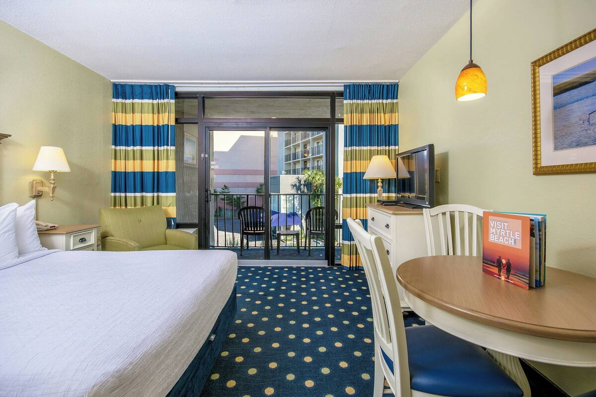 A hotel room with a bed, a desk, a chair, a table, a lamp, a framed picture, and a view of the outside balcony.