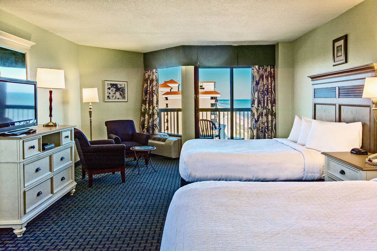A hotel room with two beds, a desk, a chair, a TV, and a view of the beach.