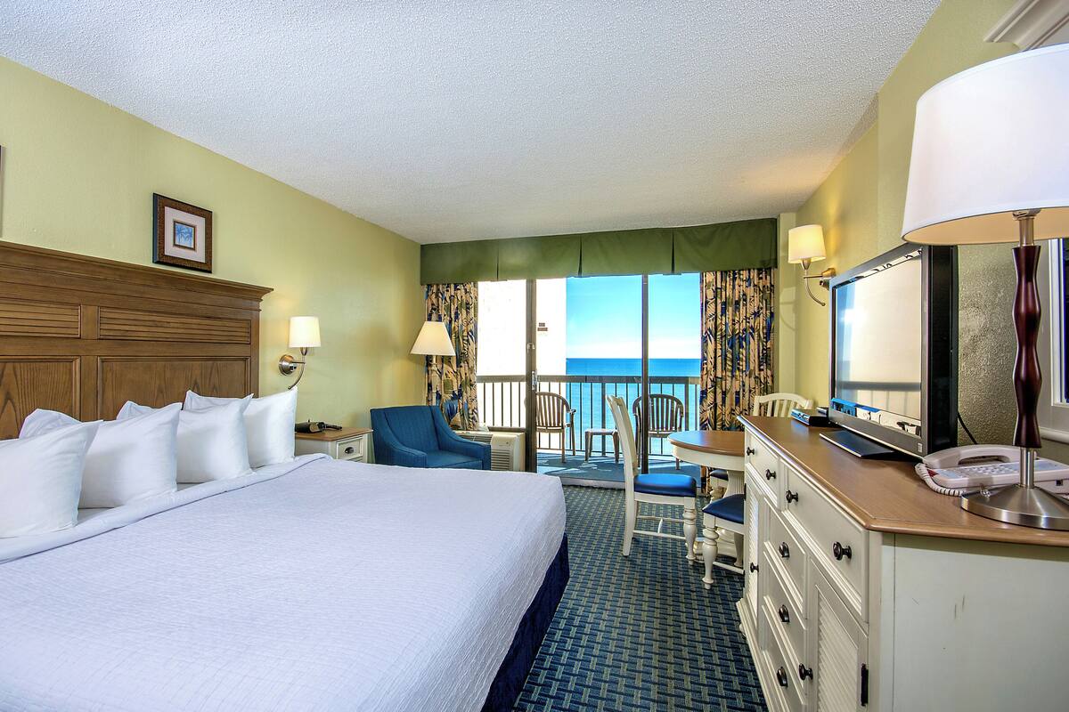 A hotel room with a large bed, a desk with a lamp, a chair, and a view of the ocean.