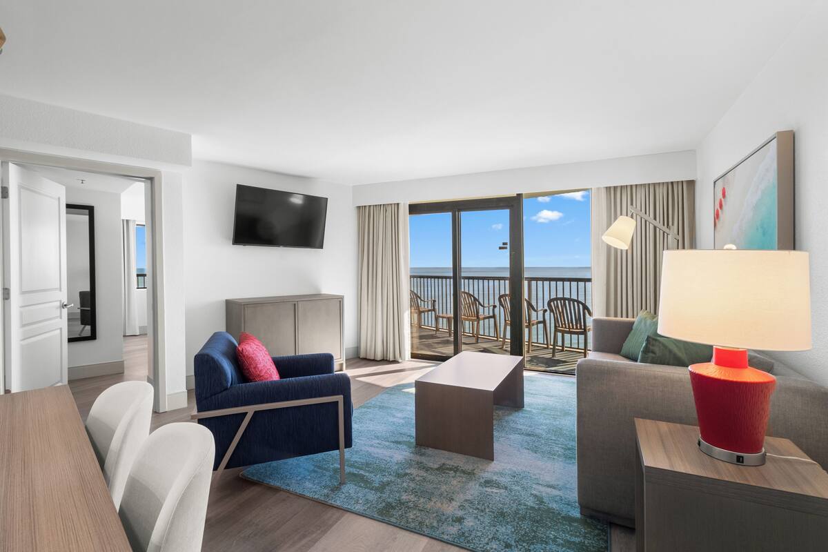 A modern hotel room with a balcony, a flat-screen TV, a sofa, a coffee table, and a lamp.