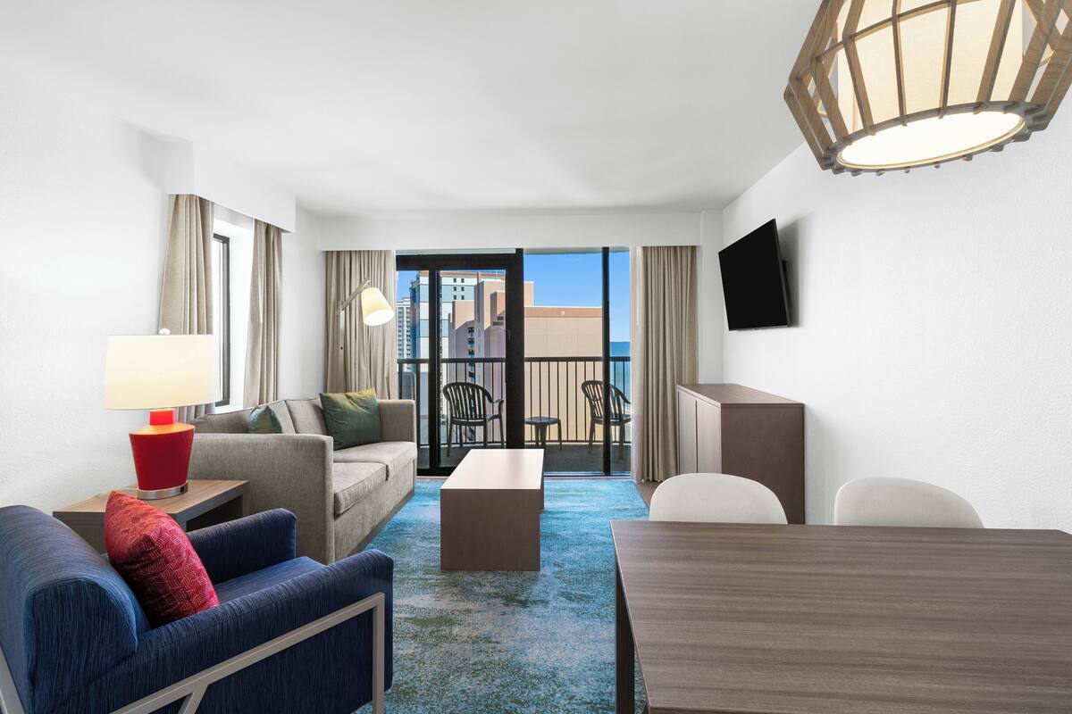 A hotel room with a sofa, armchair, a small table, a TV mounted on the wall, and a balcony with a view of the city.