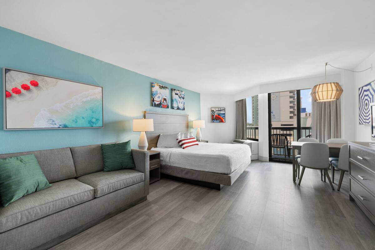 A modern hotel room with a bed, a sofa, a desk, and a balcony with a view.