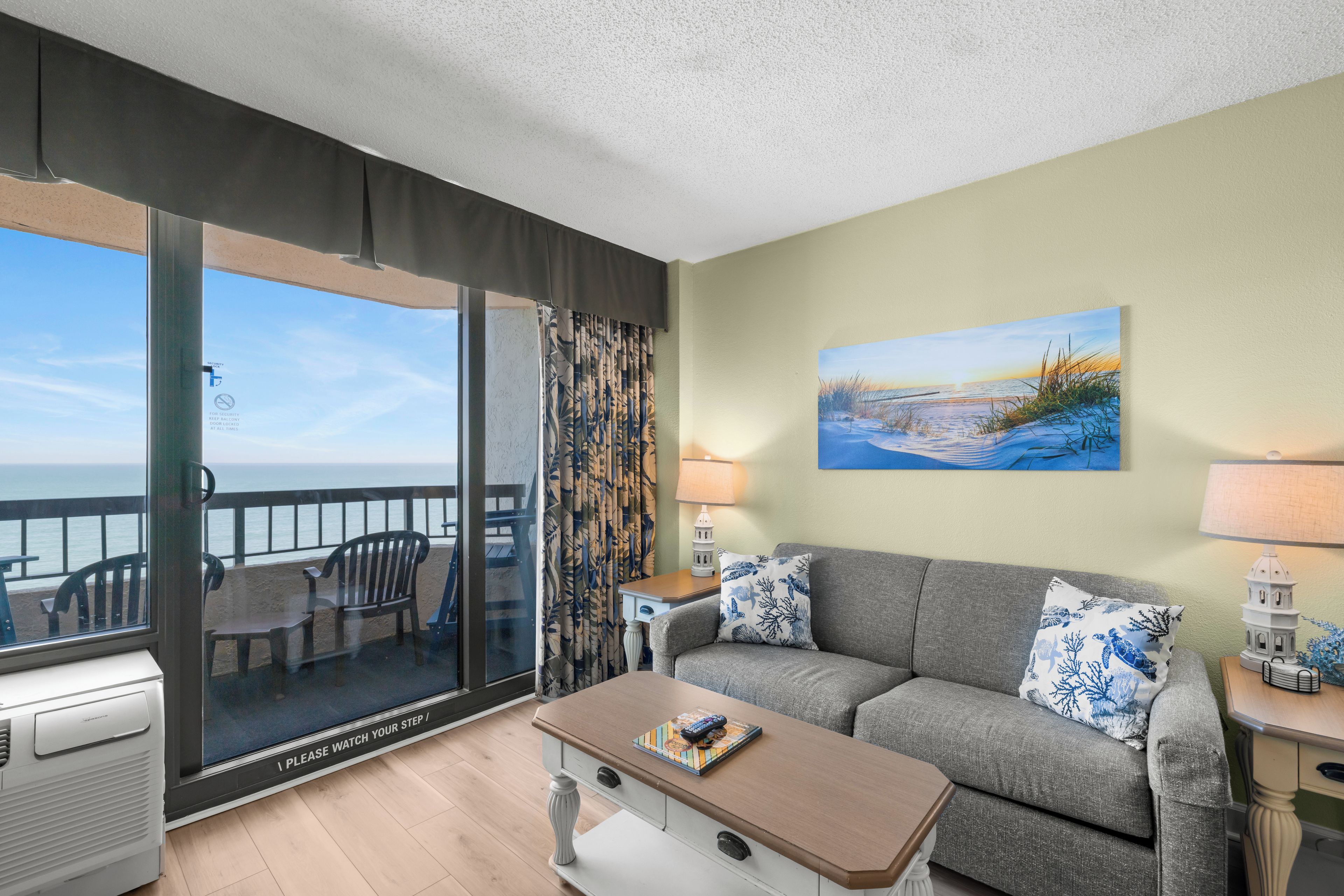 Een balkon met uitzicht op zee, een bank met decoratieve kussens, een bijzettafeltje met een spel en een schilderij aan de muur.