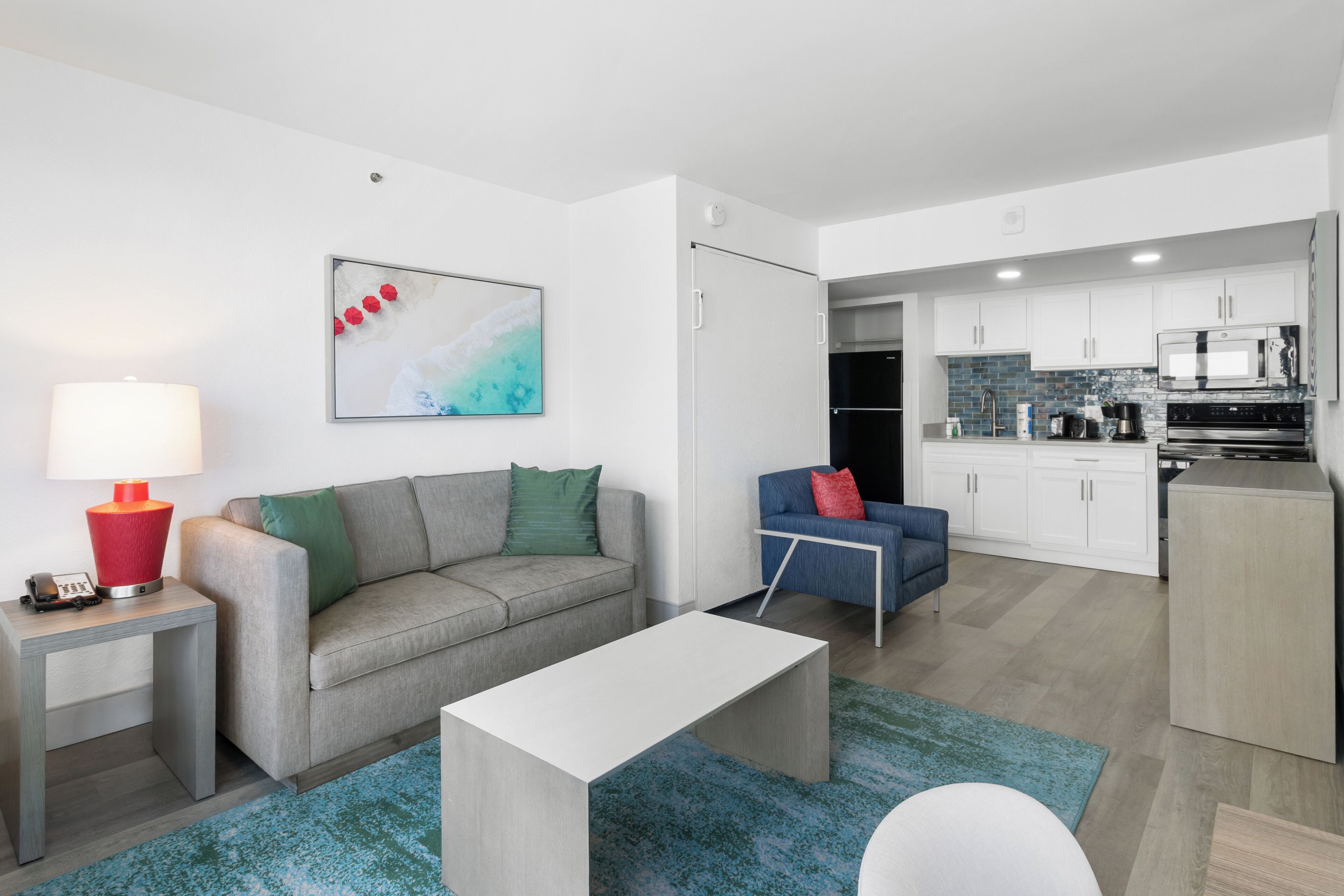 A modern living room with a grey sofa, a white coffee table, and a blue rug. There is a kitchen area with white cabinets and a microwave.