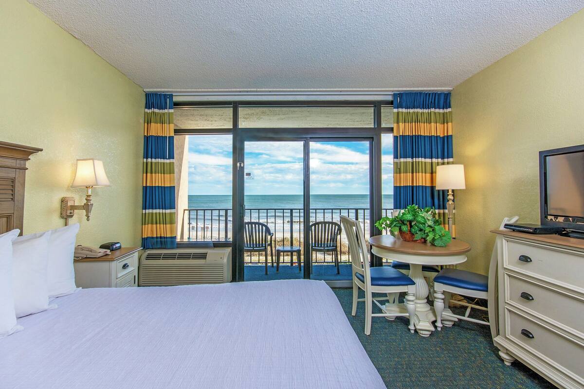 A hotel room with a bed, a desk with a chair, a television, and a view of the ocean through a sliding door.